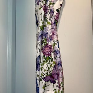 Puede incluir: Un art&iacute;culo de tela blanca con un estampado floral y de mariposas. El dise&ntilde;o presenta flores moradas y rosas, enredaderas verdes y mariposas moradas y azules. El art&iacute;culo cuelga verticalmente.