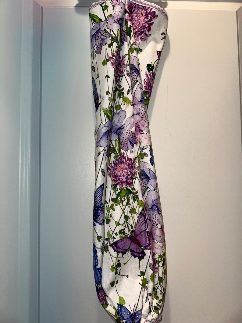 Puede incluir: Un art&iacute;culo de tela blanca con un estampado floral y de mariposas. El dise&ntilde;o presenta flores moradas y rosas, enredaderas verdes y mariposas moradas y azules. El art&iacute;culo cuelga verticalmente.