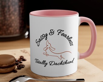 Op de afbeelding: Witte keramische koffiemok met een roze rand en handvat. De mok heeft een zwart-bruine illustratie van een teckel met de tekst "Sassy & Fearless Totally Dachshund".