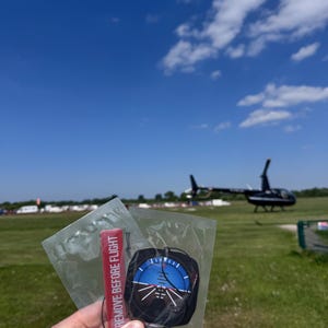 May include: A person holding a clear plastic package containing a black and blue aviation instrument and a red tag that reads "REMOVE BEFORE FLIGHT". A helicopter is visible in the background under a blue sky.