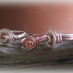 Op de afbeelding: Vijf zilveren en koperen draadringen met een spiraalvormig ontwerp. De ringen zijn in een rij op een houten oppervlak gerangschikt.