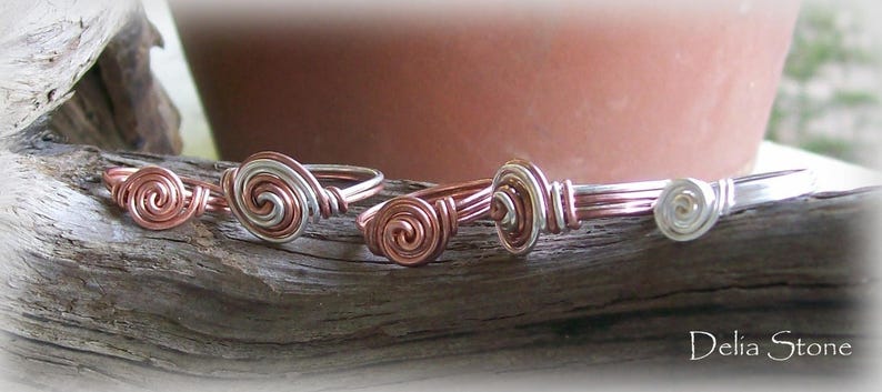 Op de afbeelding: Vijf zilveren en koperen draadringen met een spiraalvormig ontwerp. De ringen zijn in een rij op een houten oppervlak gerangschikt.