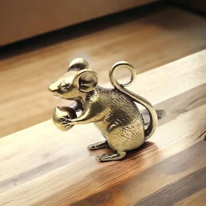 Può includere: Una statuetta di topo in ottone che tiene una noce. Il topo è dettagliato con piccole orecchie, occhi e zampe. La coda si curva verso l'alto in un anello. La statuetta è di colore dorato e poggia su una superficie di legno.