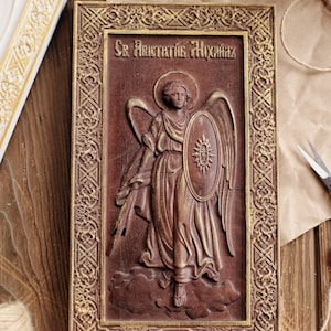 Puede incluir: Un icono de madera de San Miguel Arcángel con un marco dorado. El icono está tallado en relieve y representa al ángel con alas, una espada y un escudo. El texto "Св. Архангел Михаил" está escrito en cirílico en la parte superior del icono.