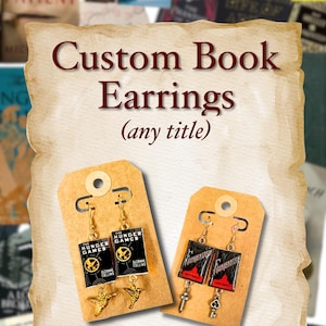 May include: Custom book earrings featuring miniature book covers. The earrings are displayed on brown cardstock with gold-colored hooks. One pair features "The Hunger Games" and the other "Frankenstein" book covers. The text "Custom Book Earrings (any title)" is displayed.
