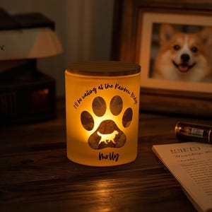 Op de afbeelding: Een witte glazen kaarsenhouder met een houten deksel. De kaarsenhouder heeft een pootprintontwerp met een hondensilhouet erin. De tekst "I'll be waiting at the Rainbow Bridge" staat gedrukt op de zijkant van de kaarsenhouder. De naam "Molly" staat gedrukt onder de pootprint.
