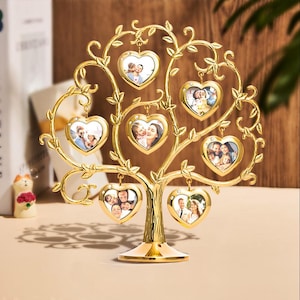 Könnte beinhalten: Ein goldfarbener Metall-Familienstammbaum-Fotorahmen. Der Baum hat herzförmige Rahmen, die an den Ästen hängen und jeweils ein Foto enthalten. Der Sockel ist rund, und der Baum hat ein dekoratives, wirbelndes Design. Der Baum ist etwa 30 cm hoch.
