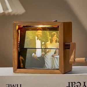 May include: A wooden photo display box with a hand crank. The box contains a photo of a couple, lit from within. The box is rectangular with a dark brown finish. The crank is made of wood and is attached to the side of the box.