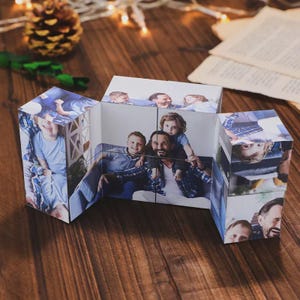 May include: A multi-sided photo cube displaying family photos. The cube is made of several connected panels, each featuring a different image of a family. The photos show a family of five, with a variety of poses and expressions. The cube is on a wooden surface.