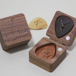 Könnte beinhalten: Holzgitarren-Plektrumboxen mit personalisierten Gravuren. Eine Box zeigt eine Gitarrenkontur und den Namen "Paul", eine andere einen Hirschkopf und den Namen "Edward". Eine dritte Box enthält ein Plektrum mit dem Buchstaben "R".