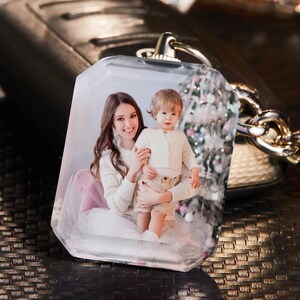 May include: A clear, rectangular keychain with a photo of a woman and a child. The photo is encased in a transparent, faceted resin. The keychain has a silver-colored metal loop and chain. The background is blurred, suggesting a close-up shot.