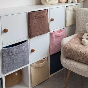 Puede incluir: Una acogedora habitación infantil con una unidad de almacenamiento blanca con contenedores de tela de varios colores. Un oso de peluche está sentado en una silla con una manta. Estantes de madera exhiben juguetes y decoración, incluyendo una cámara y arte enmarcado.