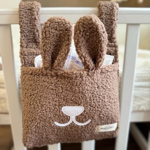 May include: A brown fuzzy bunny-shaped diaper caddy with white embroidered eyes and nose. The caddy is hanging on a white crib rail.