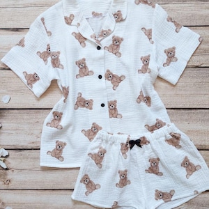 May include: White pyjama set featuring a repeating pattern of brown teddy bears. The set includes a short-sleeved button-down shirt and matching shorts with a black bow. The fabric appears to be a lightweight, textured cotton.
