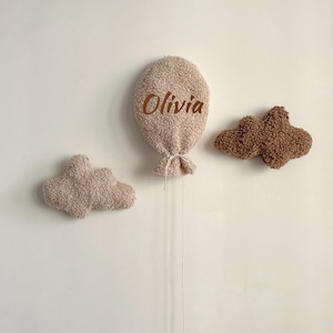 May include: A beige plush balloon with the name "Olivia" in brown script, and two cloud-shaped decorations. The balloon has a string attached. The clouds are in beige and brown.