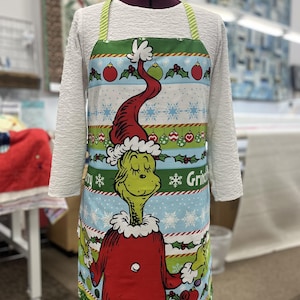 May include: A kitchen apron featuring a large graphic of a green character wearing a red Santa suit and hat. The background pattern includes horizontal stripes of light blue, white, and green with Christmas ornaments, snowflakes, and holly leaves.