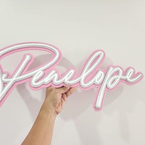 May include: Pink neon sign with the name Penelope in white script.
