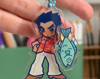 Op de afbeelding: Een doorzichtige acryl sleutelhanger met een stripfiguur met donker haar, een rood overhemd met patroon en een witte broek, die een lichtgroene vis met een eenvoudig gezicht vasthoudt. De hanger zit aan een blauwe kogelketting.