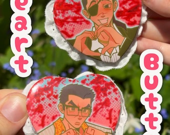 Op de afbeelding: Twee hartvormige buttons met illustraties in anime-stijl. Eén button toont een personage met een ooglapje dat met zijn handen een hart vormt. De andere button toont een personage met een bril die een duim omlaag geeft. De buttons hebben een witte kanten rand en de tekst "Heart Button" in roze.