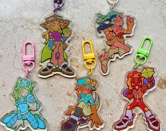 Op de afbeelding: Acryl sleutelhangers met kleurrijke anime-stijl karakterontwerpen. Elke sleutelhanger heeft een ander karakter met unieke outfits en poses. De sleutelhangers hebben verschillende gekleurde sluitingen, waaronder paars, roze, geel en groen.