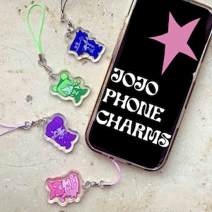 Puede incluir: Cuatro colgantes de acrílico para teléfono con ilustraciones de personajes estilizadas en azul, verde, morado y rosa, cada uno con un cordón a juego. Un smartphone muestra una estrella rosa y el texto JOJO PHONE CHARMS en blanco.