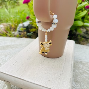 May include: A white beaded charm bracelet with a gold-tone Highland cow charm. The bracelet is on a white wooden stand.