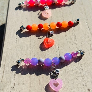 May include: Three colorful beaded bracelets with heart-shaped charms. Each charm has a letter on it, spelling out the name "J.R.". The bracelets are pink, orange, and purple.