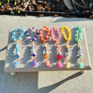 May include: Seven colorful beaded bracelets with gummy bear charms. Each bracelet features a different color and pattern of beads, including white, blue, purple, orange, pink, yellow, and green. The bracelets are displayed on a wooden surface.