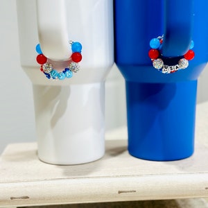May include: Two white mugs with blue handles, one white and one blue, with red, white, and blue beaded bracelets that spell out "USA" around the handles.