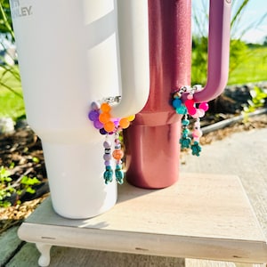 May include: Two stainless steel tumblers with colorful beaded charms attached to the handles. The tumblers are on a white wooden stand. The charms feature small turquoise turtle figurines.