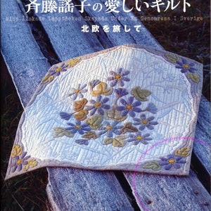 May include: A white quilt with a floral design featuring blue and green flowers. The quilt is folded and resting on a wooden surface. The text "斉藤謠子の愛しいキルト" is written in Japanese at the top of the image.