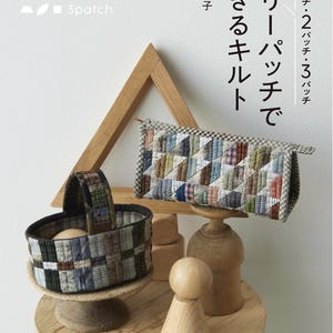 Puede incluir: Un libro titulado "スリーパッチでできるキルト" (Suri Patchi dekiru Kiruto) con una cesta y una bolsa acolchadas. La cesta y la bolsa están hechas de tela de patchwork en varios colores. También hay objetos decorativos de madera.