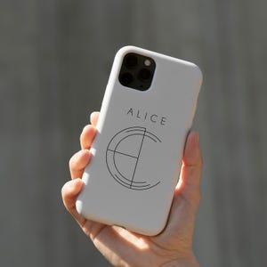 May include: A white phone case with the name "ALICE" printed in black, along with a geometric design. The case is held in a hand, showcasing the design and the phone's camera array.