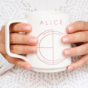 May include: White ceramic mug with the name "ALICE" in burgundy lettering and a matching geometric design. The mug is held by two hands. The mug has a large handle.
