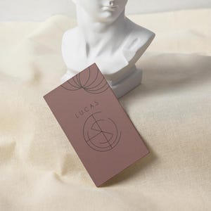 May include: A rectangular business card in a muted mauve color, featuring the name "LUCAS" in a simple font and a circular graphic design. The card is positioned in front of a white bust sculpture on a cream-colored fabric.