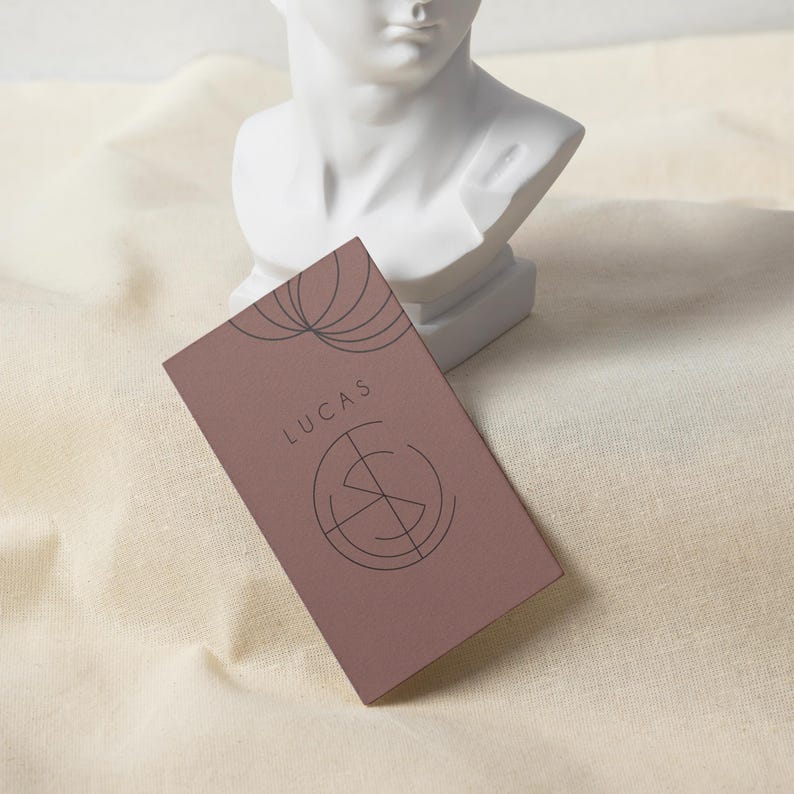 May include: A rectangular business card in a muted mauve color, featuring the name "LUCAS" in a simple font and a circular graphic design. The card is positioned in front of a white bust sculpture on a cream-colored fabric.