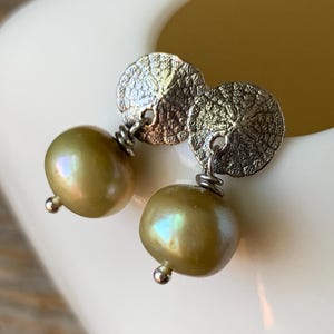 May include: A pair of earrings featuring olive-green, iridescent pearls suspended from textured silver-toned circular studs. The pearls have a subtle sheen, and the earrings are set against a white background.