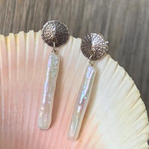 May include: A pair of dangle earrings featuring textured silver-tone disc studs and long, iridescent white freshwater pearls. The earrings are displayed on a seashell against a wooden background. The pearls have a rectangular shape.