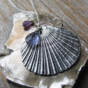 May include: A silver seashell pendant necklace with two purple teardrop-shaped gemstones. The necklace is displayed on a piece of rough, gray rock.