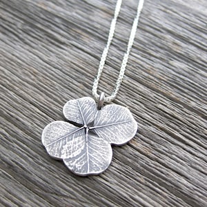 May include: A silver necklace with a four-leaf clover pendant. The pendant is detailed with a textured design that resembles the veins of a leaf.