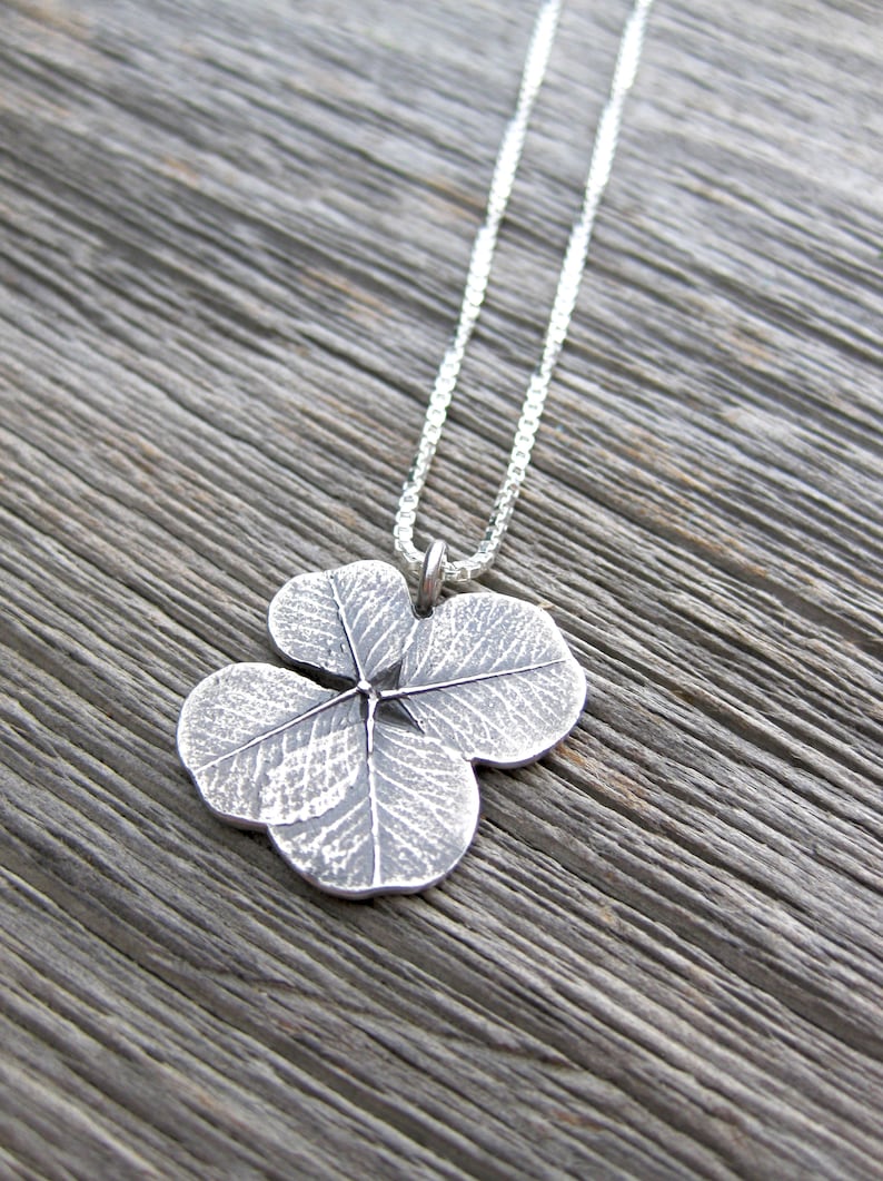 May include: A silver necklace with a four-leaf clover pendant. The pendant is detailed with a textured design that resembles the veins of a leaf.