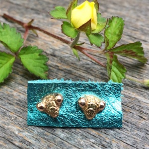 May include: A pair of gold-toned stud earrings with a textured, triangular design. The earrings are displayed on a turquoise leather background.