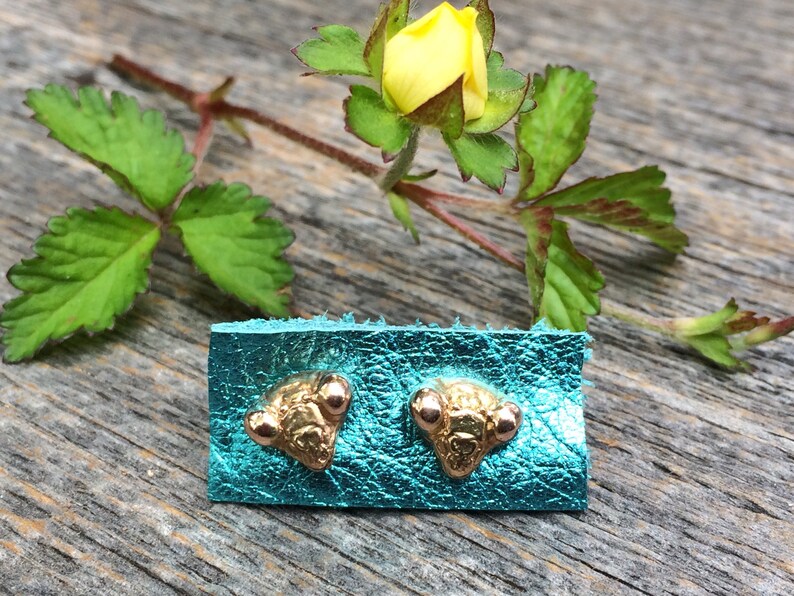 May include: A pair of gold-toned stud earrings with a textured, triangular design. The earrings are displayed on a turquoise leather background.