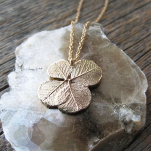 May include: A gold necklace with a four-leaf clover pendant. The pendant is detailed with textured lines and is on a light brown background.