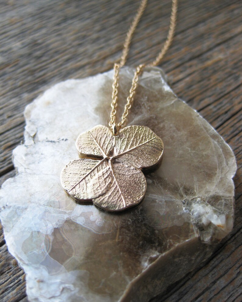 May include: A gold necklace with a four-leaf clover pendant. The pendant is detailed with textured lines and is on a light brown background.
