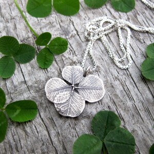 May include: A silver four-leaf clover pendant necklace on a silver chain. The pendant is detailed with a textured design that resembles the veins of a leaf. The necklace is displayed on a wooden surface with green four-leaf clovers.