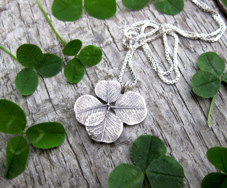 May include: A silver four-leaf clover pendant necklace on a silver chain. The pendant is detailed with a textured design that resembles the veins of a leaf. The necklace is displayed on a wooden surface with green four-leaf clovers.