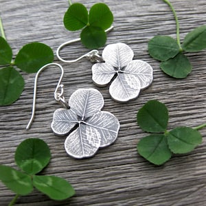 May include: A pair of silver earrings shaped like four-leaf clovers. The earrings are hanging from silver hooks and are set against a wooden background with green four-leaf clovers.
