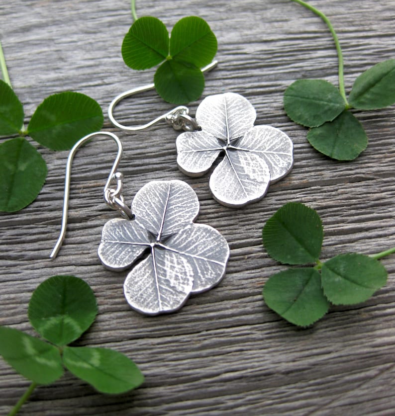 May include: A pair of silver earrings shaped like four-leaf clovers. The earrings are hanging from silver hooks and are set against a wooden background with green four-leaf clovers.