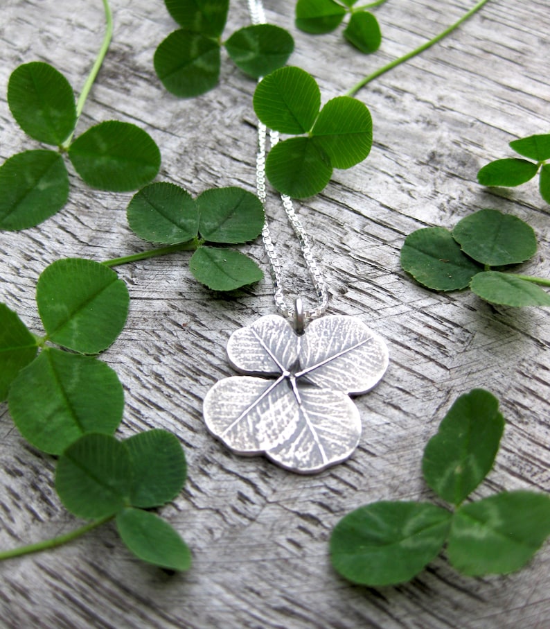 May include: A silver four-leaf clover pendant necklace on a silver chain. The necklace is lying on a wooden surface with green four-leaf clover leaves scattered around it.